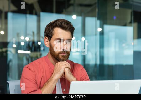 Close-up photo. Concentrated and thoughtful young man in a red shirt works, studies on a laptop in the office Stock Photo