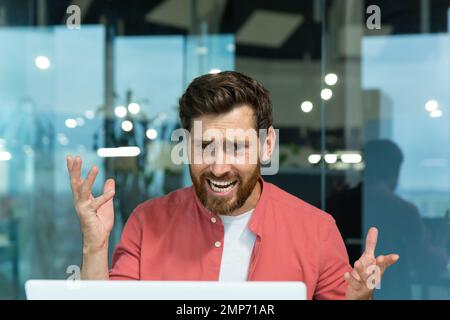 Close up photo. An angry young man in a red shirt is sitting in the office and talking on a video call from a laptop. Screams, aggressively gestures with his hands, explains. Stock Photo