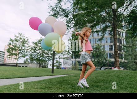 Little blond girl posing with baloons in city and looking at camera. Stock Photo