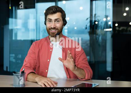 A young man in a red shirt in a headset sits in the office at the table and conducts business training on camera. Explains, advises, smiles. Stock Photo