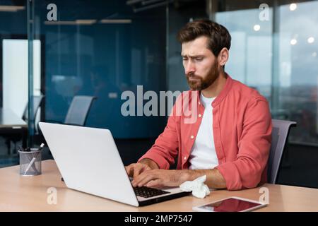 Tired young male designer in a red shirt works in the office at a laptop. He feels bad, got sick, uses a napkin. Stock Photo
