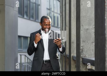Happy young african american man standing near office center in suit. He holds the phone in his hands, looks at the display, celebrates, rejoices. I received good news, messages, winnings. Stock Photo
