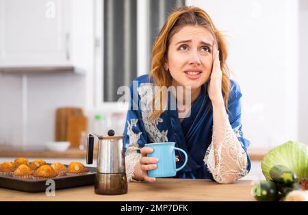 Sleepy woman in housecoat drinking morning coffee in kitchen Stock Photo