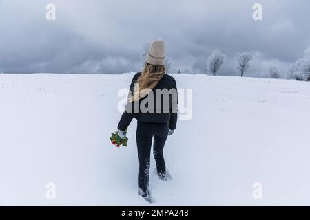 Young hiker woman in a black jacket with a bouquet of flowers in her hands walks in the snow. Stock Photo