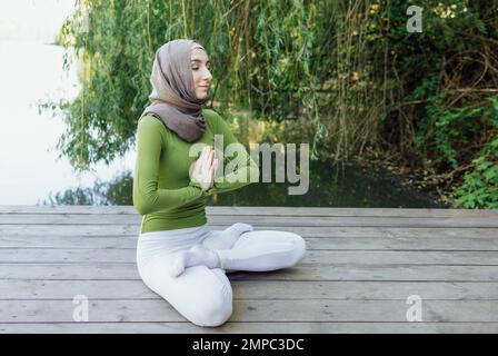 Muslim young girl in a hijab is doing yoga asana in the park. Teenage girl sits in a lotus position on pier by the lake or river. Relax concept Stock Photo