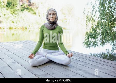 Muslim young girl in a hijab is doing yoga asana in the park. Teenage girl sits in a lotus position on pier by the lake or river. Relax concept Stock Photo