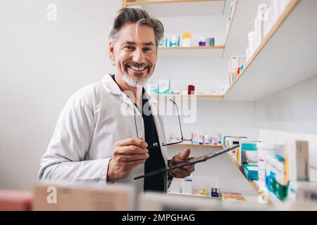 Senior pharmacist standing in a chemist and looking at the camera. Mature healthcare worker holding a digital tablet and eyeglasses in a pharmacy. Stock Photo