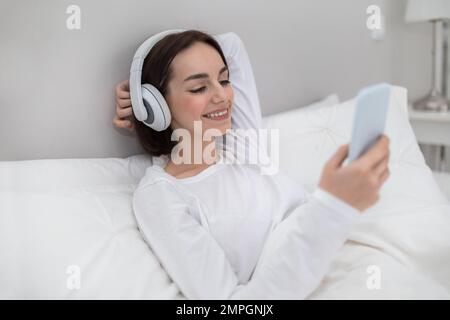 Cheery young woman chilling in bed with phone and headphones Stock Photo