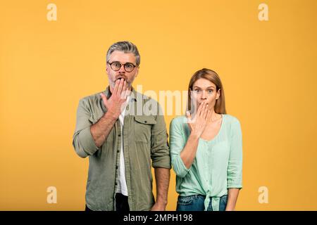Shocked middle aged caucasian spouses covering mouths in shock and looking at camera, standing on yellow background Stock Photo