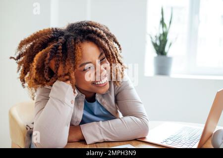 Young African American woman is working on a laptop. Focused female student is looking at computer screen and watching webinar or doing video chat by Stock Photo