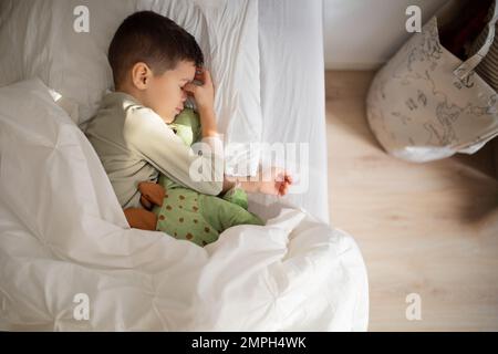 Tired european small kid in pajamas sleeps, wakes up on bed with dinosaur toy in bedroom interior Stock Photo