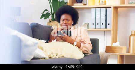Sick and tired woman lying on a couch typing on her phone at home. A young African female relaxing and yawing on a sofa in her house on a boring Stock Photo