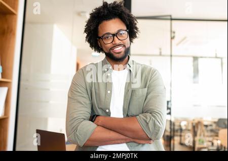 Portrait of confident male office employee in stylish glasses and casual shirt, smart and ambitious african-american man with curly hair looks at the camera with hands crossed Stock Photo