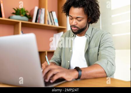 Focused and concentrated african-american male student, office worker using laptop for web searching, web surfing. Curly purposeful man in casual shirt making notes in notebook, researching Stock Photo