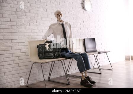 Human skeleton in office wear sitting on chair near brick wall indoors Stock Photo