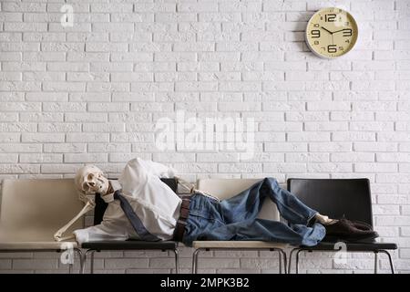 Human skeleton in office wear lying on chairs near brick wall indoors Stock Photo