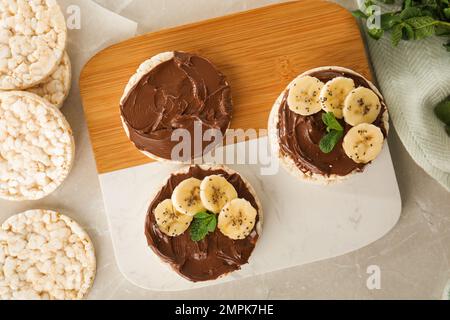 Puffed rice cakes with chocolate spread, banana and mint on grey marble table, flat lay Stock Photo