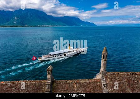 View over Lake Geneva and alpine scenery in the distance over the roofs of Chillon Castle, Château de Chillon, a vintage steam boat passing by. Stock Photo