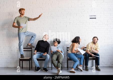 redhead man standing on chair and rehearsing near multiethnic actors sitting and waiting for casting Stock Photo