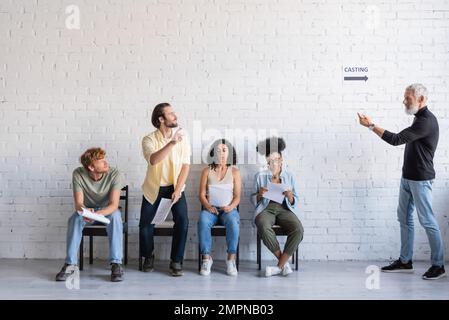bearded art director calling brunette man near multiethnic actors sitting on chairs and waiting for casting Stock Photo