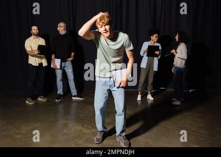 confused redhead man holding clipboard and touching head near multiethnic students and teacher of acting skills Stock Photo