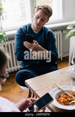 Young man using smart phone sitting at dining table in home Stock Photo
