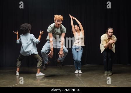 excited redhead man jumping near multiethnic actors posing during rehearsal in theater Stock Photo