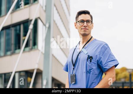 Confident male doctor wearing blue scrubs looking away in front of hospital building Stock Photo