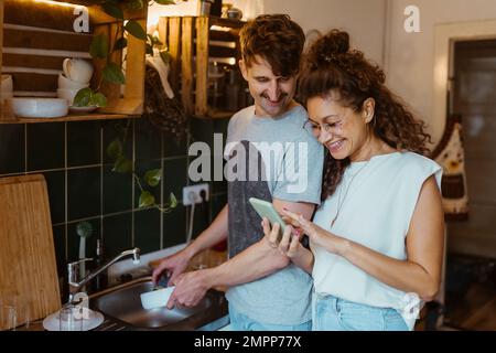 Smiling man doing dishes while looking at smart phone held by girlfriend in kitchen Stock Photo