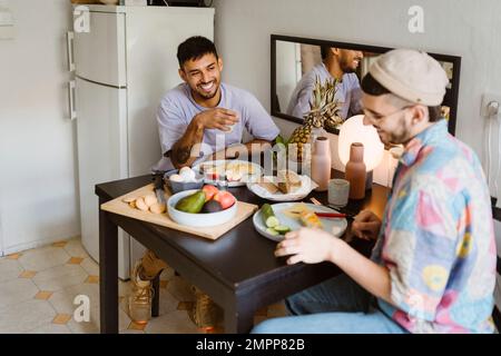 Happy gay couple having fruits while sitting at dining table in kitchen Stock Photo