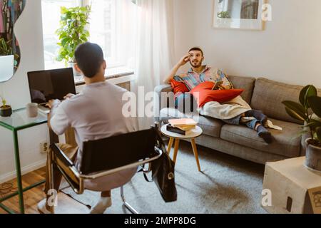 Man on sofa talking to boyfriend sitting with laptop at home Stock Photo