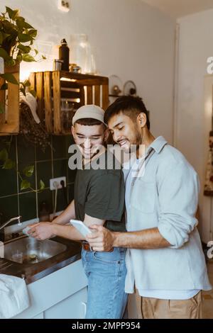 Smiling man washing dishes while boyfriend sharing smart phone in kitchen at home Stock Photo