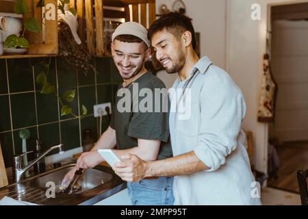 Smiling man sharing smart phone with boyfriend doing dishes in kitchen at home Stock Photo