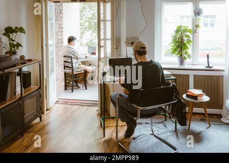 Rear view of gay couple using laptops while sitting on chairs at home Stock Photo