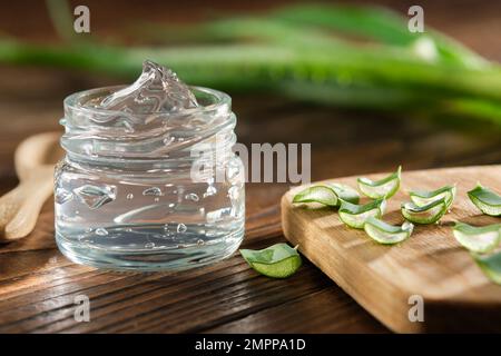 Transparent glass jar of aloe vera gel, sliced aloe leaves on a cutting board. Aloe vera plant on background. Natural cosmetic and medicinal remedy. Stock Photo