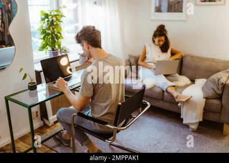 Man having coffee while using laptop sitting on chair near girlfriend in background at home Stock Photo