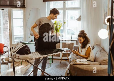 Man ironing clothes looking at woman using smart phone on sofa at home Stock Photo