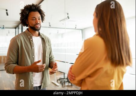 Two coworkers chatting during coffee break standing in glass hall of office building. Curly african-american male employee explain something for female colleague, sharing his ideas Stock Photo