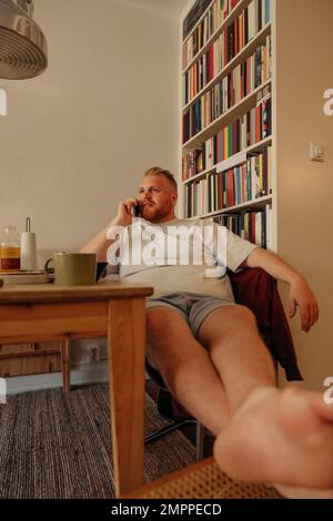 Man talking on mobile phone while sitting near bookshelf at home Stock Photo
