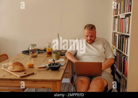 Man using laptop while sitting with breakfast by bookshelf at home Stock Photo