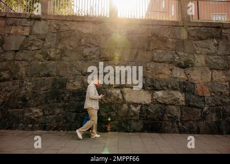 Man using smart phone while walking on footpath in front of wall Stock Photo