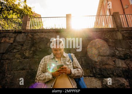 Low angle view of smiling man using smart phone in front of wall during sunny day Stock Photo
