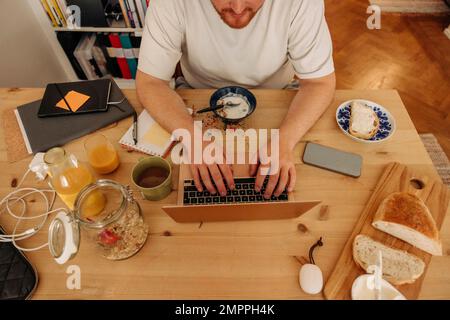 High angle view of man typing on laptop while sitting with breakfast on table at home Stock Photo