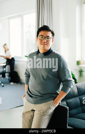 Portrait of smiling young businessman with hands in pockets leaning on sofa at office Stock Photo