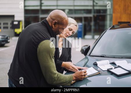 Saleswoman assisting male customer while doing paperwork standing near car Stock Photo