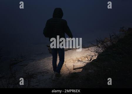 Man with flashlight walking near river in evening Stock Photo