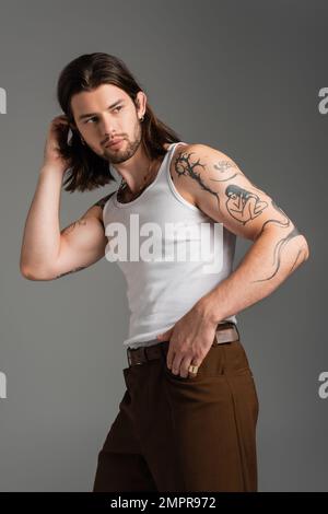 Trendy tattooed man in white sleeveless shirt adjusting hair isolated on grey Stock Photo