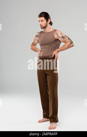 Barefoot and tattooed man in pants and tank top posing on grey background Stock Photo