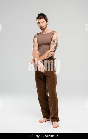 Full length of young barefoot and tattooed model looking at camera on grey background Stock Photo