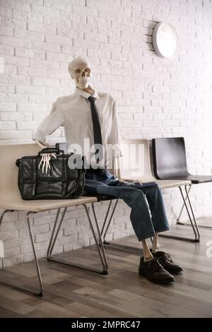 Human skeleton in office wear sitting on chair near brick wall indoors Stock Photo